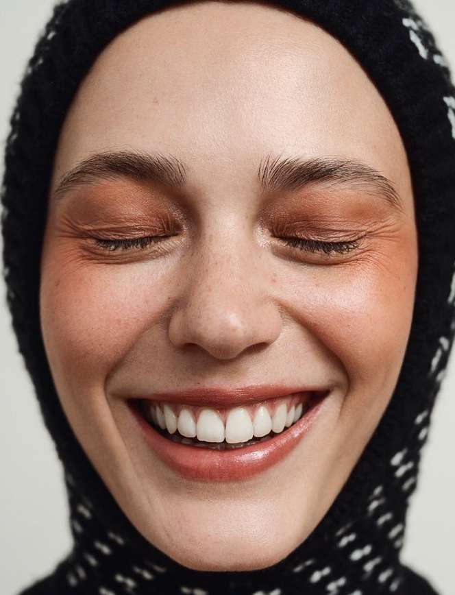 Close-up portrait of a young adult woman smiling with her eyes closed, wearing natural makeup and a black and white patterned knit hat.