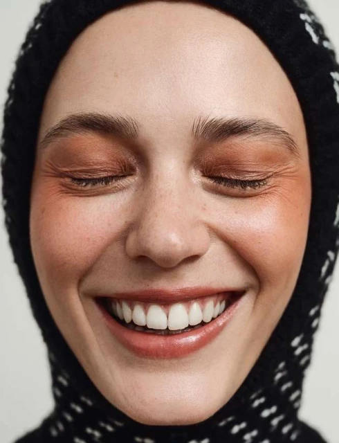 Close-up portrait of a young adult woman smiling with her eyes closed, wearing natural makeup and a black and white patterned knit hat.