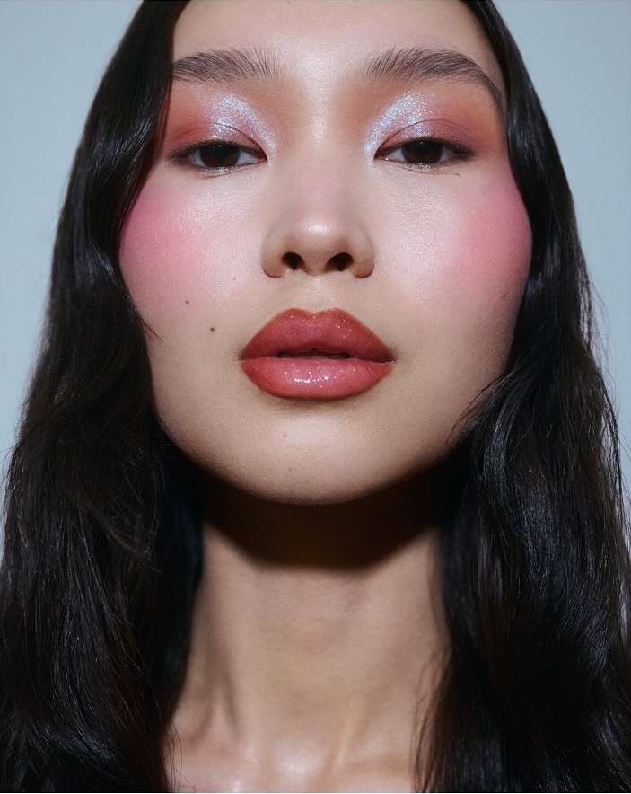 Close-up portrait of an adult female with dark wavy hair and glamorous pink and orange makeup in an indoor setting with a neutral background.