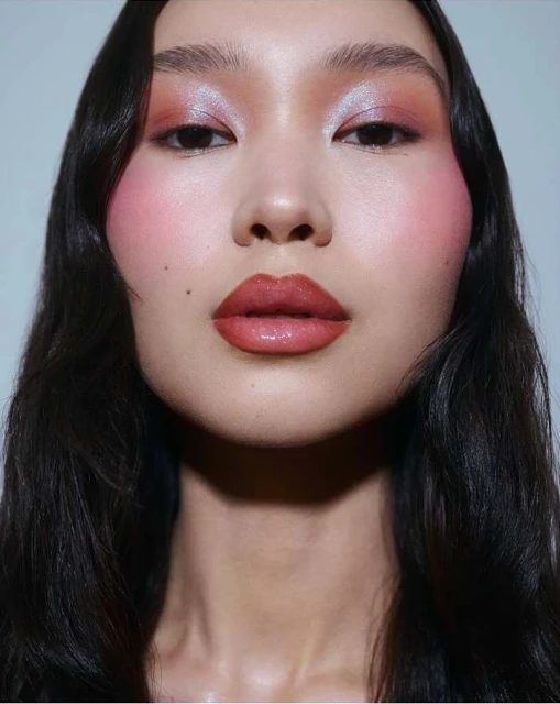 Close-up portrait of an adult female with dark wavy hair and glamorous pink and orange makeup in an indoor setting with a neutral background.