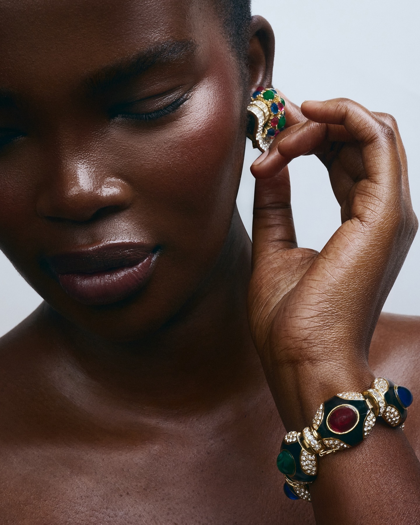Close-up of a dark-skinned woman showcasing statement jewelry, including gold earrings and a bracelet with red, green, and blue gemstones.