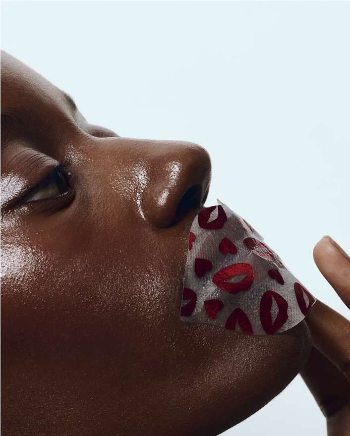 Close-up of a Black woman's face with a patterned lip mask featuring red lips and hearts, against a stark white background.