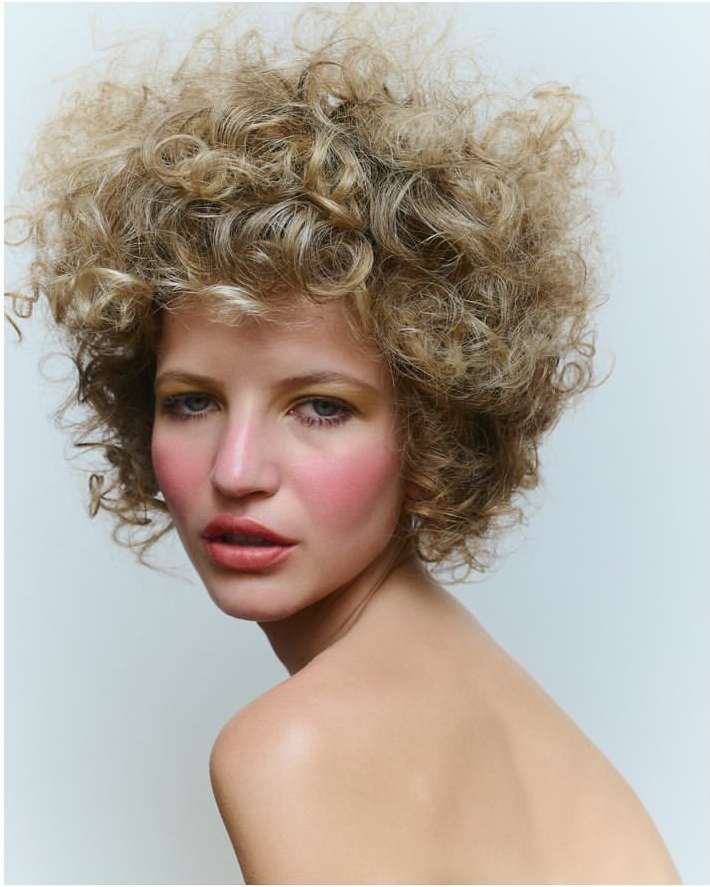 Close-up portrait of an adult woman with curly blonde hair, wearing makeup, in a studio setting with a light-colored background.