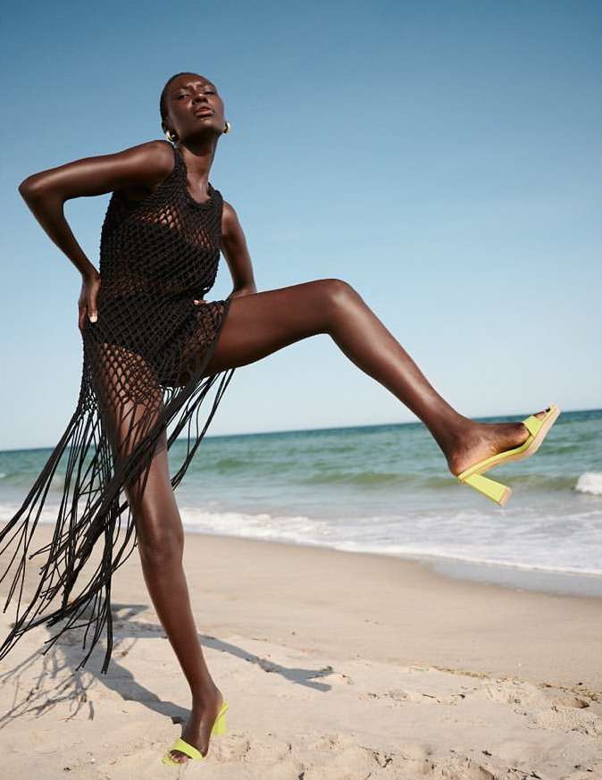 Woman in black cover-up and neon heels poses on the beach against blue sky and ocean backdrop, in fashion and lifestyle shot.