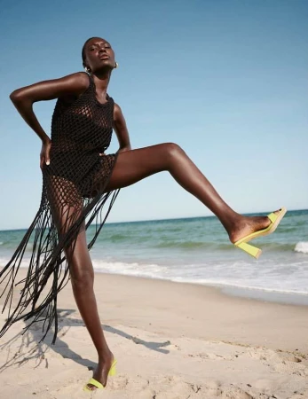 Woman in black cover-up and neon heels poses on the beach against blue sky and ocean backdrop, in fashion and lifestyle shot.