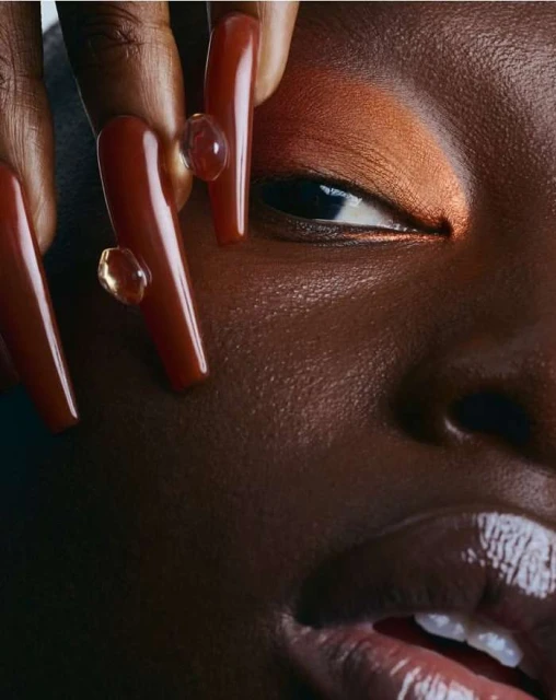 Close-up shot of an adult woman with dramatic bronze eye makeup, long brown nails with beads, and dark skin in a beauty-style photo.
