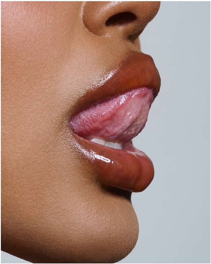 Close-up side profile of an adult female with medium skin tone, licking her lips, which are glossed with caramel-colored lip gloss.