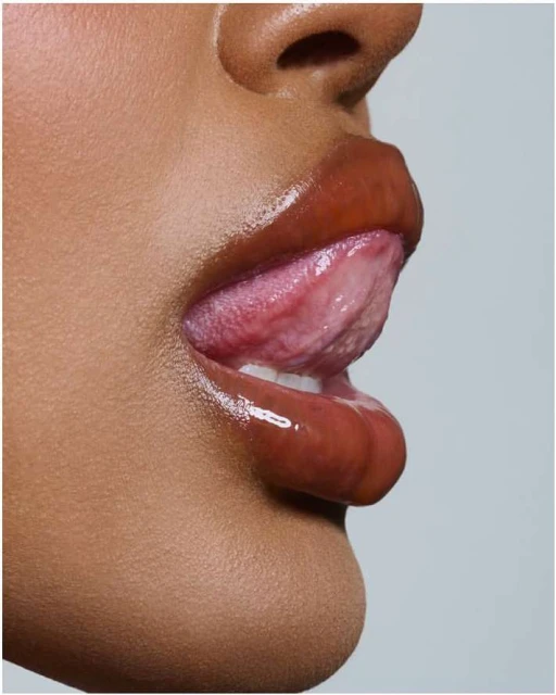 Close-up side profile of an adult female with medium skin tone, licking her lips, which are glossed with caramel-colored lip gloss.
