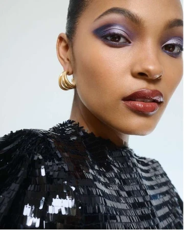 Close-up portrait of a Black woman with dramatic purple eye shadow wearing a black, sequined top and gold hoop earrings.