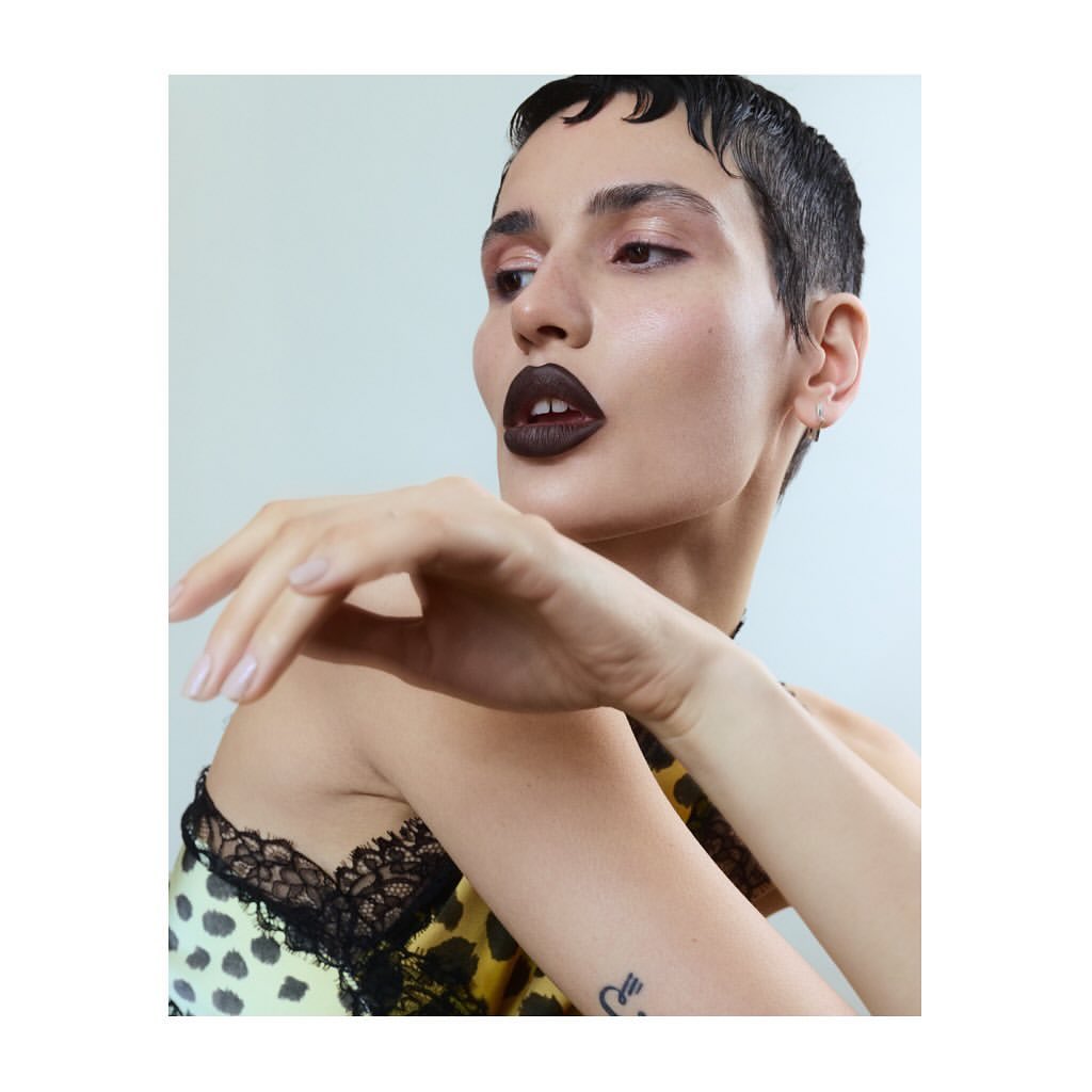 Close-up portrait of a young woman with dark lipstick and a yellow animal-print top, set against a neutral background in a beauty editorial style.