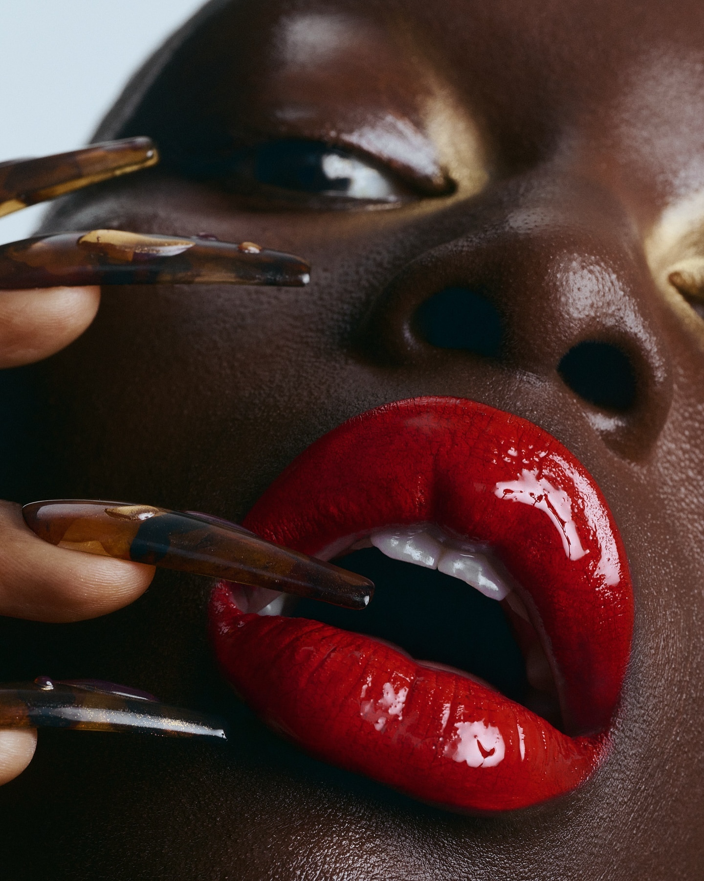 Close-up of a dark-skinned adult female face with red lips and long nails. Beauty shot shows face, red lipstick, brown skin, and long brown nails.