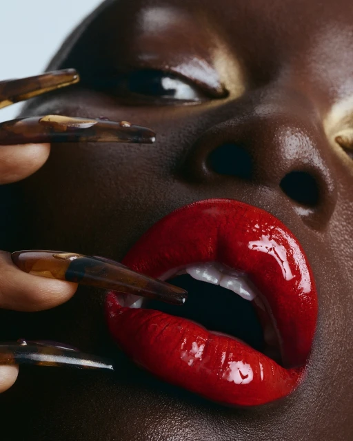Close-up of a dark-skinned adult female face with red lips and long nails. Beauty shot shows face, red lipstick, brown skin, and long brown nails.