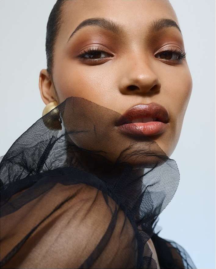 Close-up studio shot of a model with a natural makeup look wearing a sheer black ruffled top against a white backdrop.