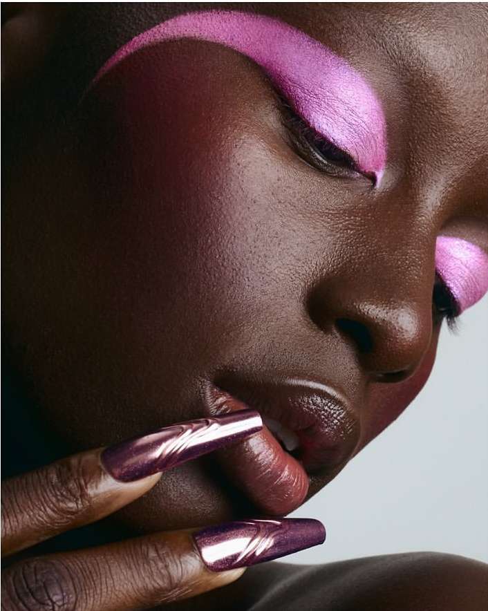 Close-up of a Black woman with long, metallic purple nails and glossy pink makeup on her eyelids and eyebrows against a light background.