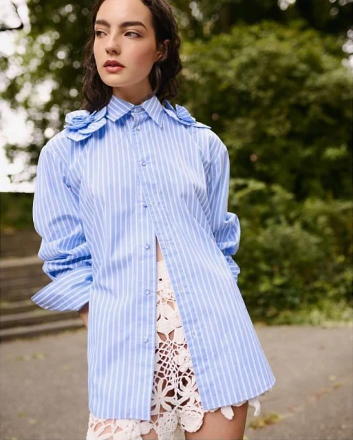 Elegant woman in an open striped shirt and crochet shorts stands outdoors against a soft-focus green tree background in natural light.