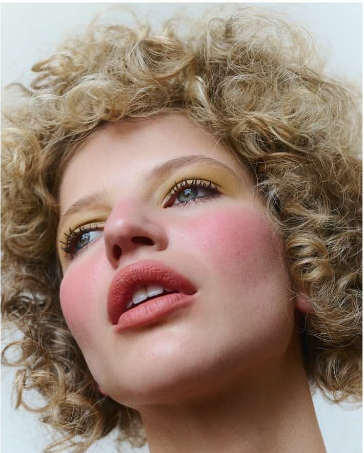 Close-up of a young adult woman with curly blonde hair and make-up featuring yellow eyeshadow and pink blush, angled low against a white backdrop.