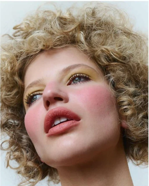 Close-up of a young adult woman with curly blonde hair and make-up featuring yellow eyeshadow and pink blush, angled low against a white backdrop.