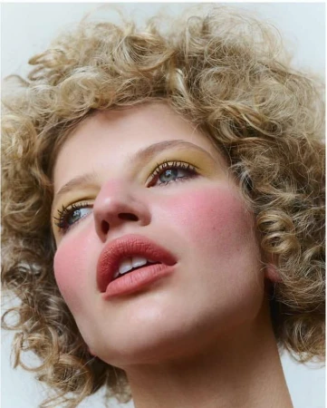 Close-up of a young adult woman with curly blonde hair and make-up featuring yellow eyeshadow and pink blush, angled low against a white backdrop.