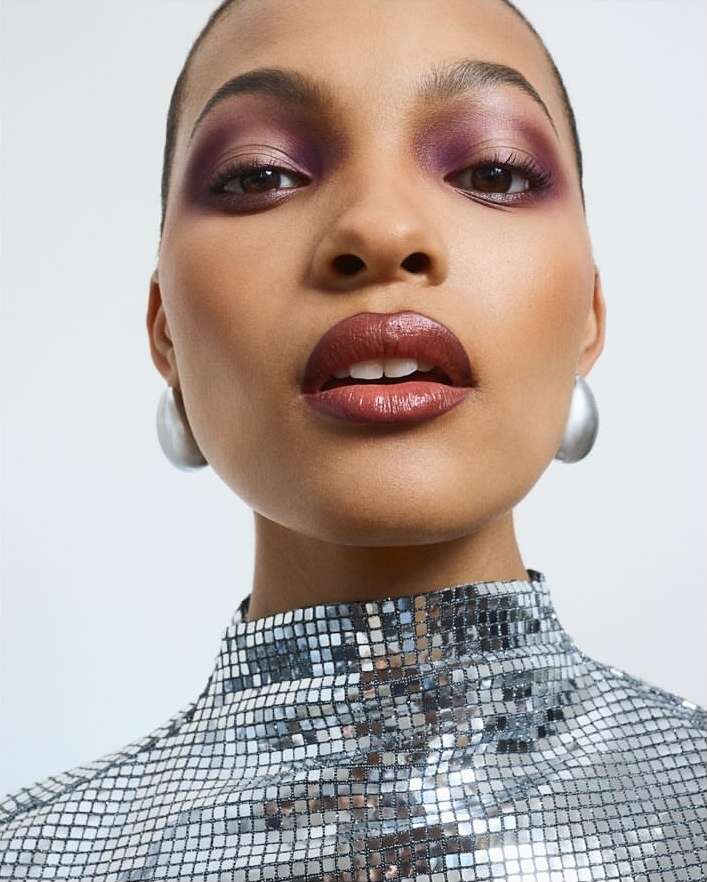 Close-up portrait of a young woman with dramatic purple eyeshadow, glossy lips, and silver earrings wearing a reflective, square-patterned top.