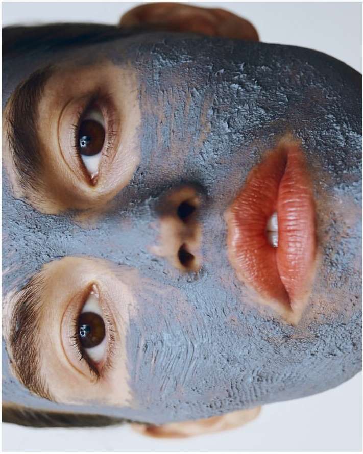 Close-up of an adult female face with a gray facial mask. The model's eyes are open, and lips are red, shot against a white background.
