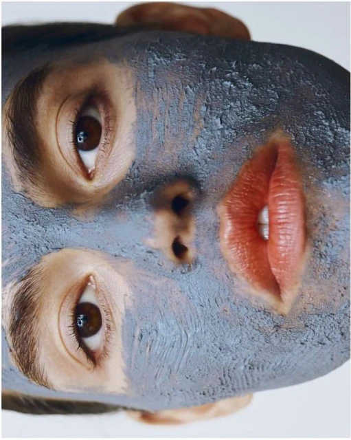 Close-up of an adult female face with a gray facial mask. The model's eyes are open, and lips are red, shot against a white background.
