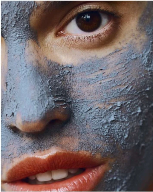 Close-up of a woman's face with a grey facial mask, light skin, brown eyes, orange lips, and white teeth, showcasing a beauty treatment.