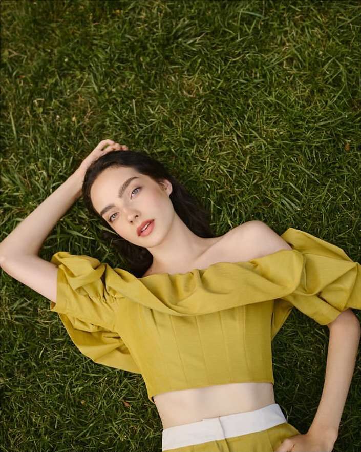 A woman lying down on green grass wearing an off-the-shoulder yellow top, captured from a high-angle overhead perspective.