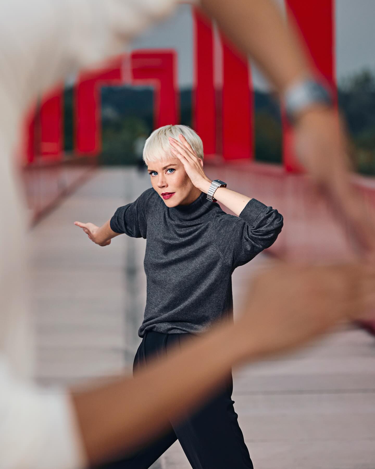 Woman with short blond hair in front of a red bridge with a person slightly out of focus in the foreground. Woman with arm raised.