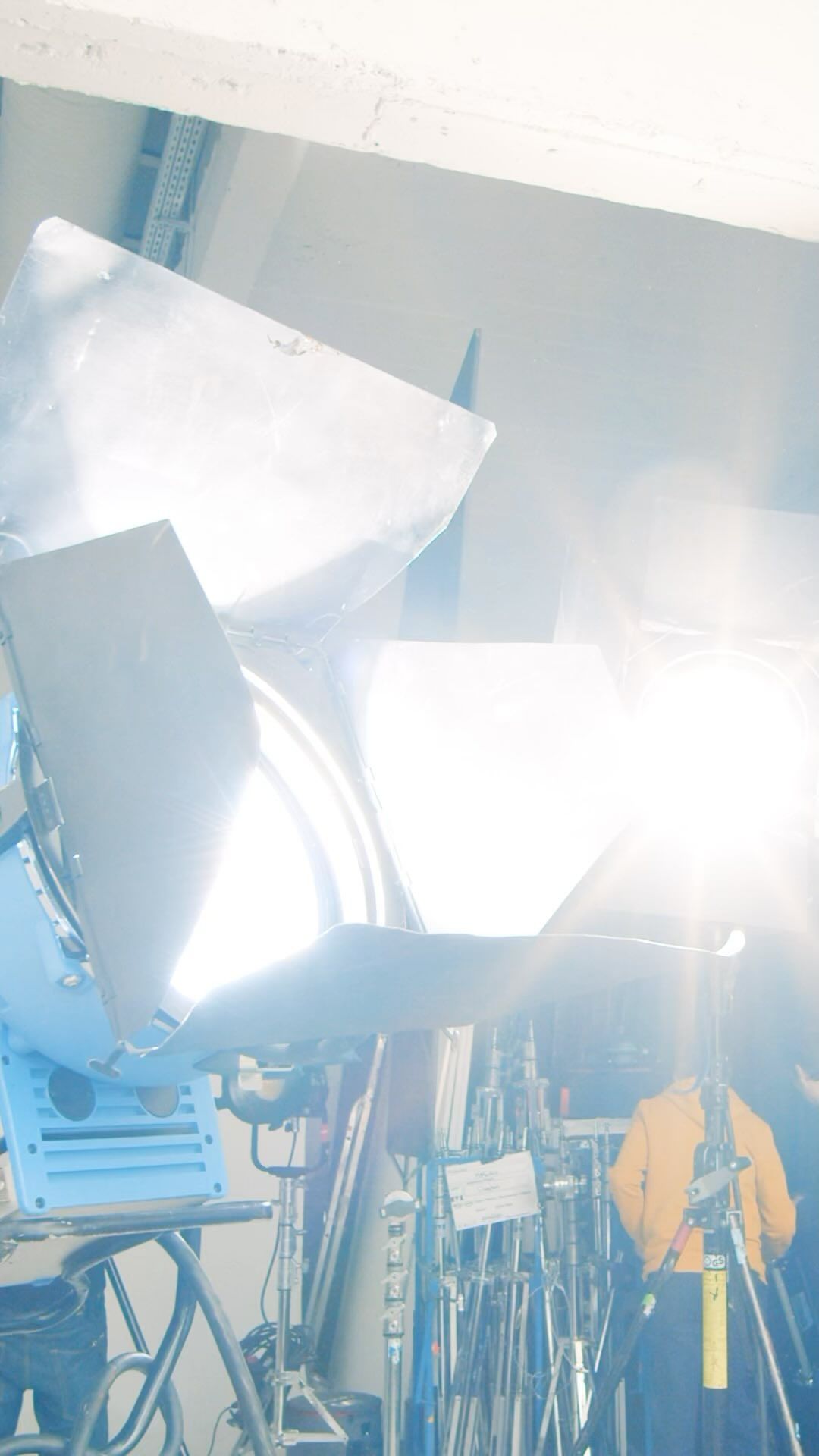 Illuminated film set with bright spotlights, scaffolding, stands and one person wearing a yellow jacket in the background.