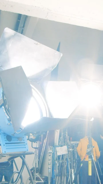 Illuminated film set with bright spotlights, scaffolding, stands and one person wearing a yellow jacket in the background.