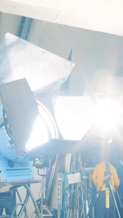 Illuminated film set with bright spotlights, scaffolding, stands and one person wearing a yellow jacket in the background.