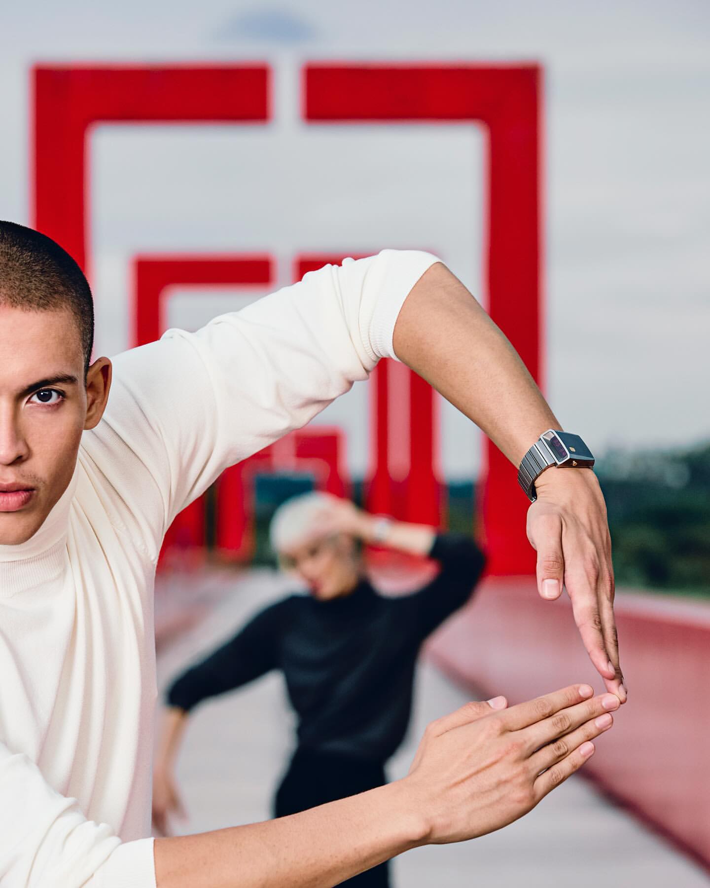 Fashion shot featuring a man in a white turtleneck with a watch, striking a pose, with a woman in black in the background.