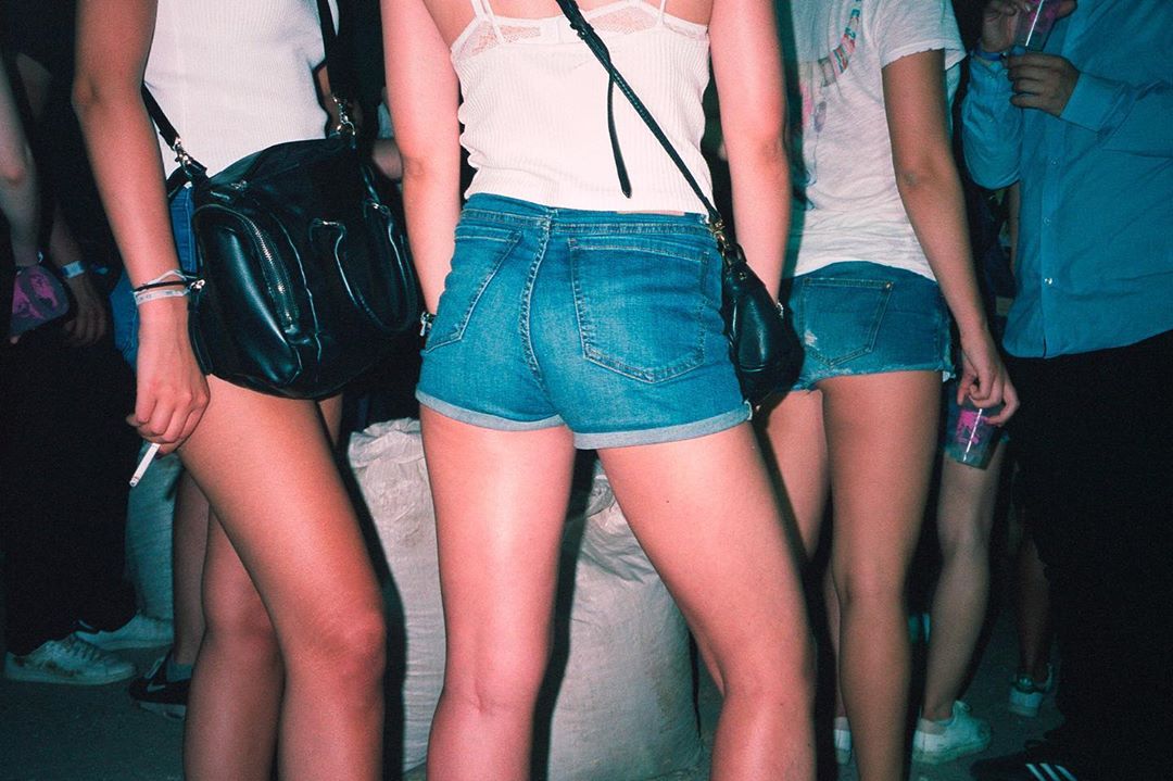 A group of young adult women wearing denim shorts and tank tops, standing together in a dimly lit indoor setting with dark background.
