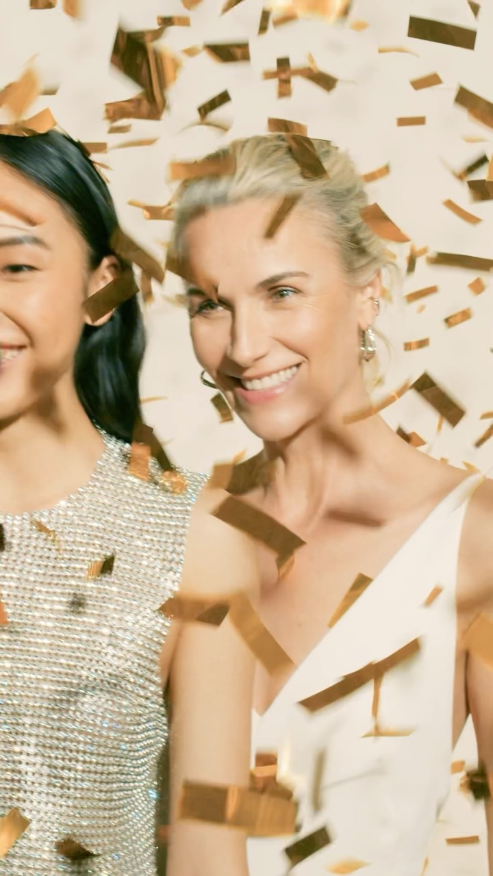 Two smiling women, one Asian and one white, stand indoors in front of a neutral background with gold confetti falling on them.