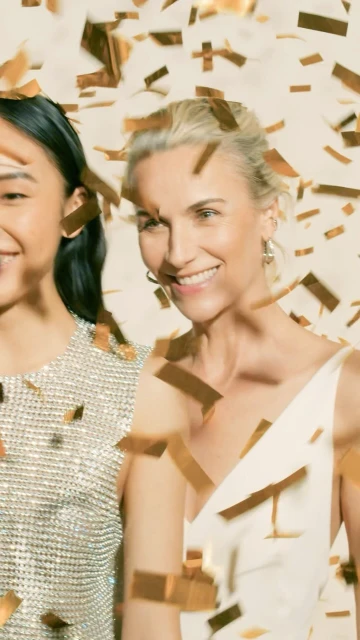 Two smiling women, one Asian and one white, stand indoors in front of a neutral background with gold confetti falling on them.