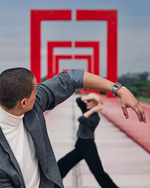 Two adults pose in front of red geometric arches, with one man facing away, showing off a metallic watch, and the other dancing in the background.
