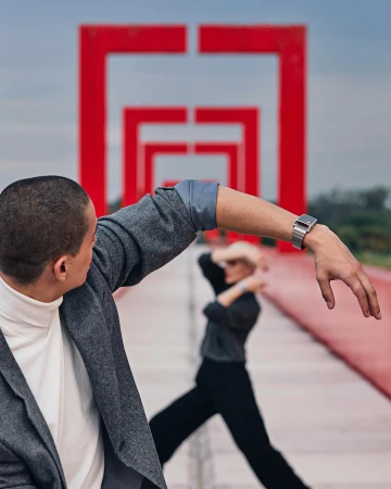 Two adults pose in front of red geometric arches, with one man facing away, showing off a metallic watch, and the other dancing in the background.