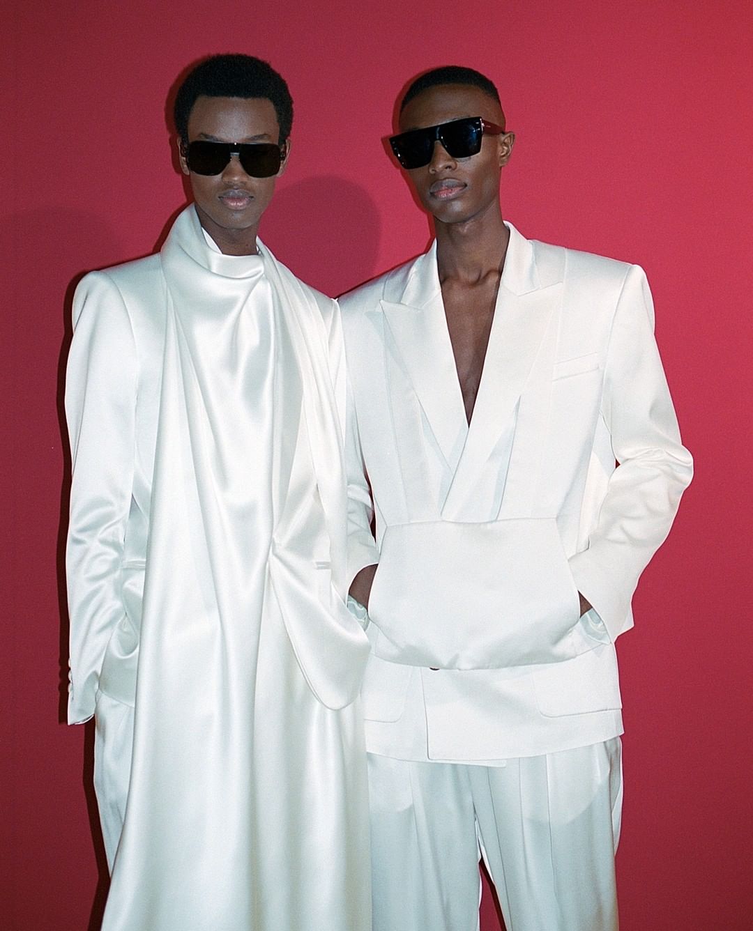 Two dark-skinned male fashion models wearing sunglasses and white matching suits in front of a simple red background in a studio.