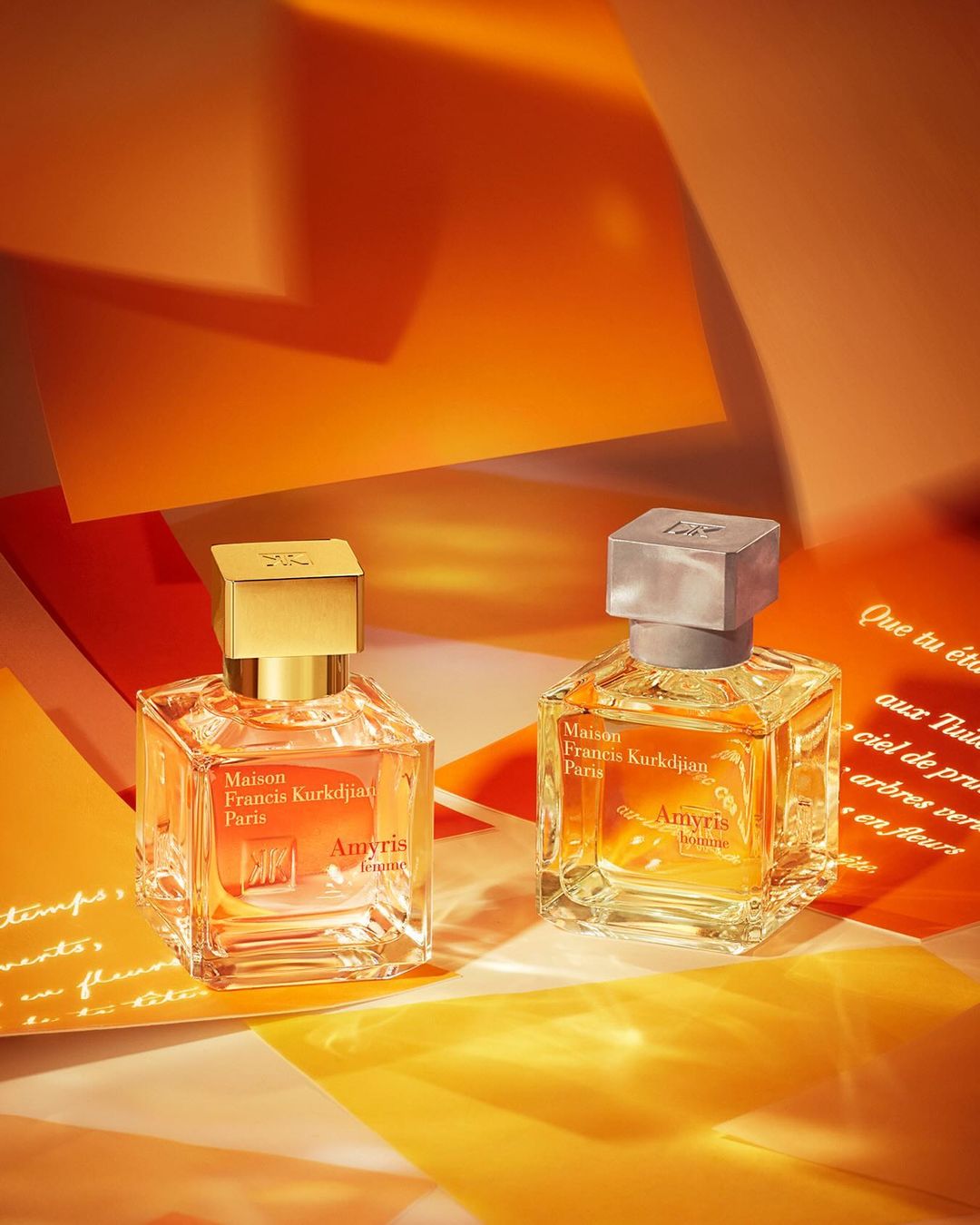 Two square glass perfume bottles, one with a gold cap and the other with a silver cap, are placed on yellow and orange paper.