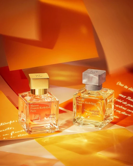 Two square glass perfume bottles, one with a gold cap and the other with a silver cap, are placed on yellow and orange paper.