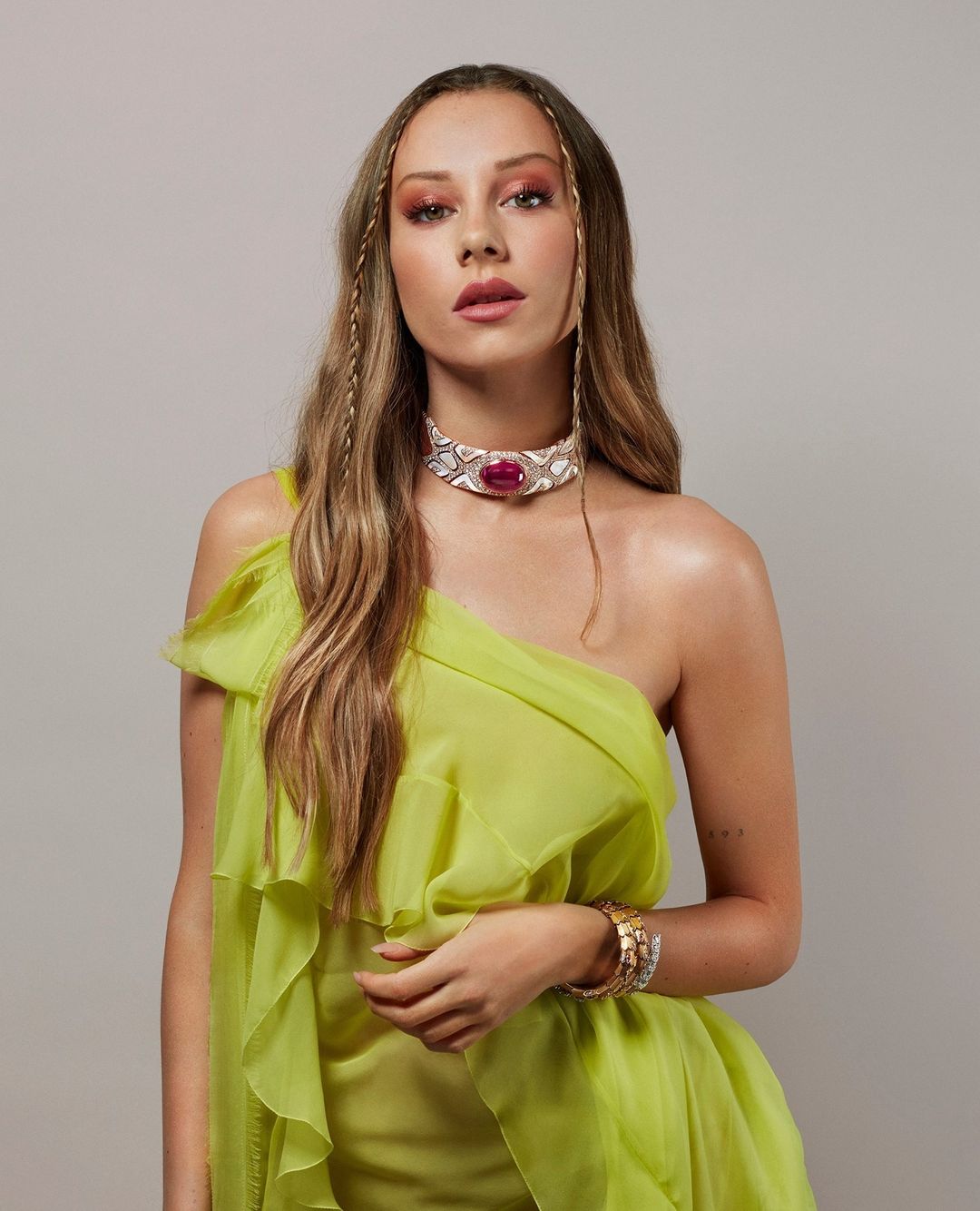 Portrait of a blonde woman wearing a ruffled neon-yellow one-shoulder dress and jeweled choker against a grey background.
