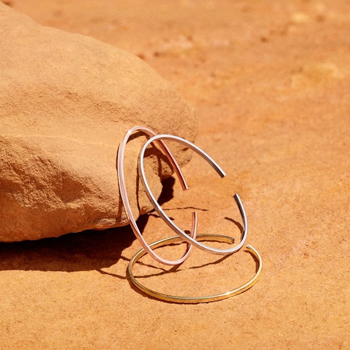 Close-up of three open-ended metal bracelets (gold, silver, rose gold) arranged on a textured sandstone surface under bright, diffused sunlight.