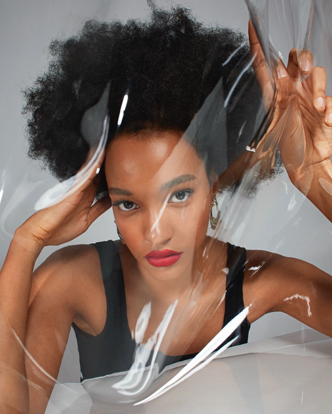 Portrait of a young woman with afro hairstyle and red lipstick poses with hands near head, partially obscured by a clear plastic sheet.