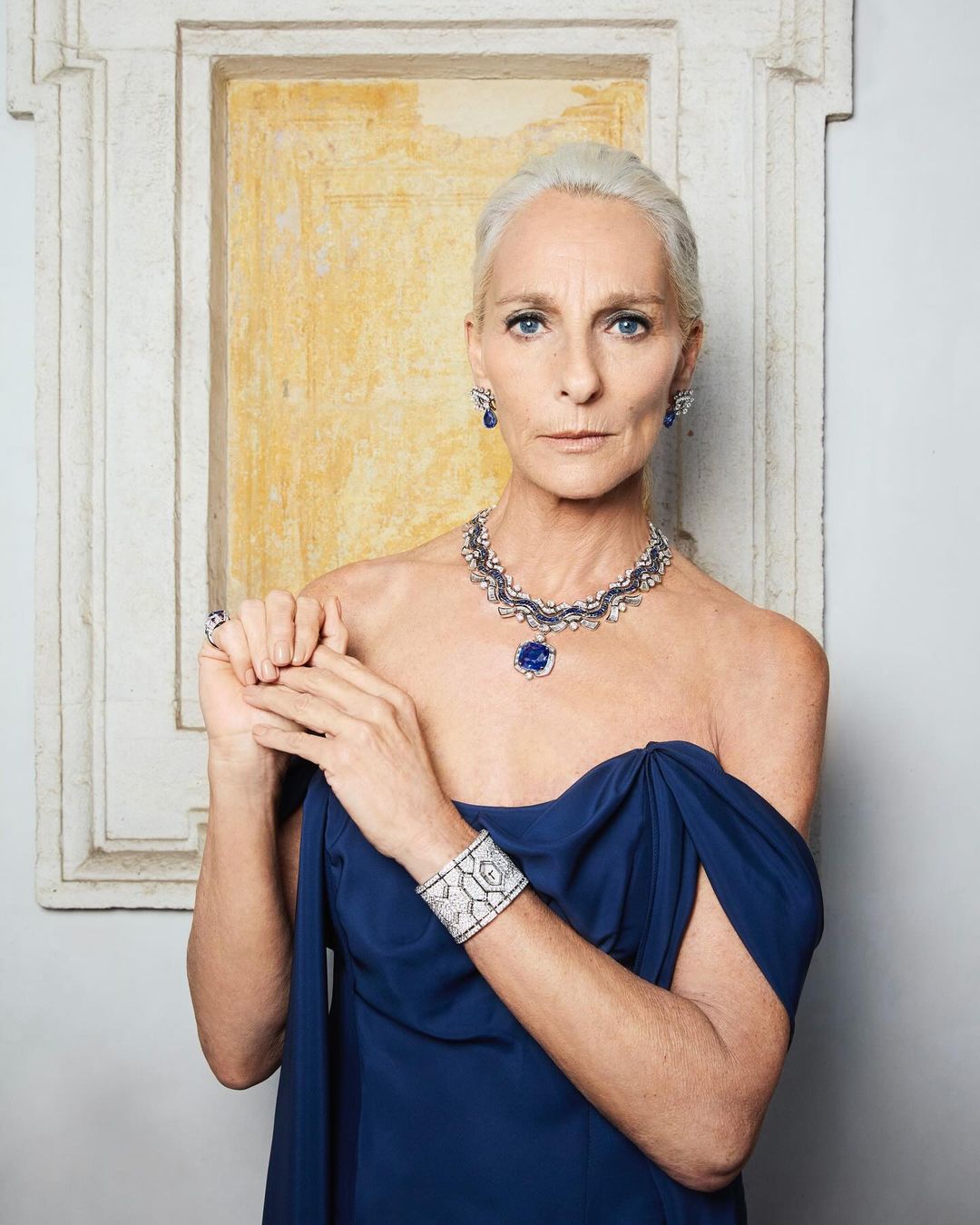 Elegant older woman poses wearing an off-the-shoulder blue dress and diamond and sapphire jewelry in front of a decorative wall.