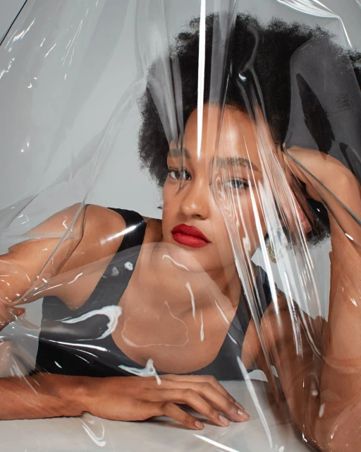 Close-up portrait of a woman with dark curly hair and red lipstick posing behind a translucent plastic sheet in a photo studio.