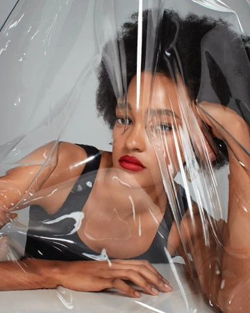 Close-up portrait of a woman with dark curly hair and red lipstick posing behind a translucent plastic sheet in a photo studio.