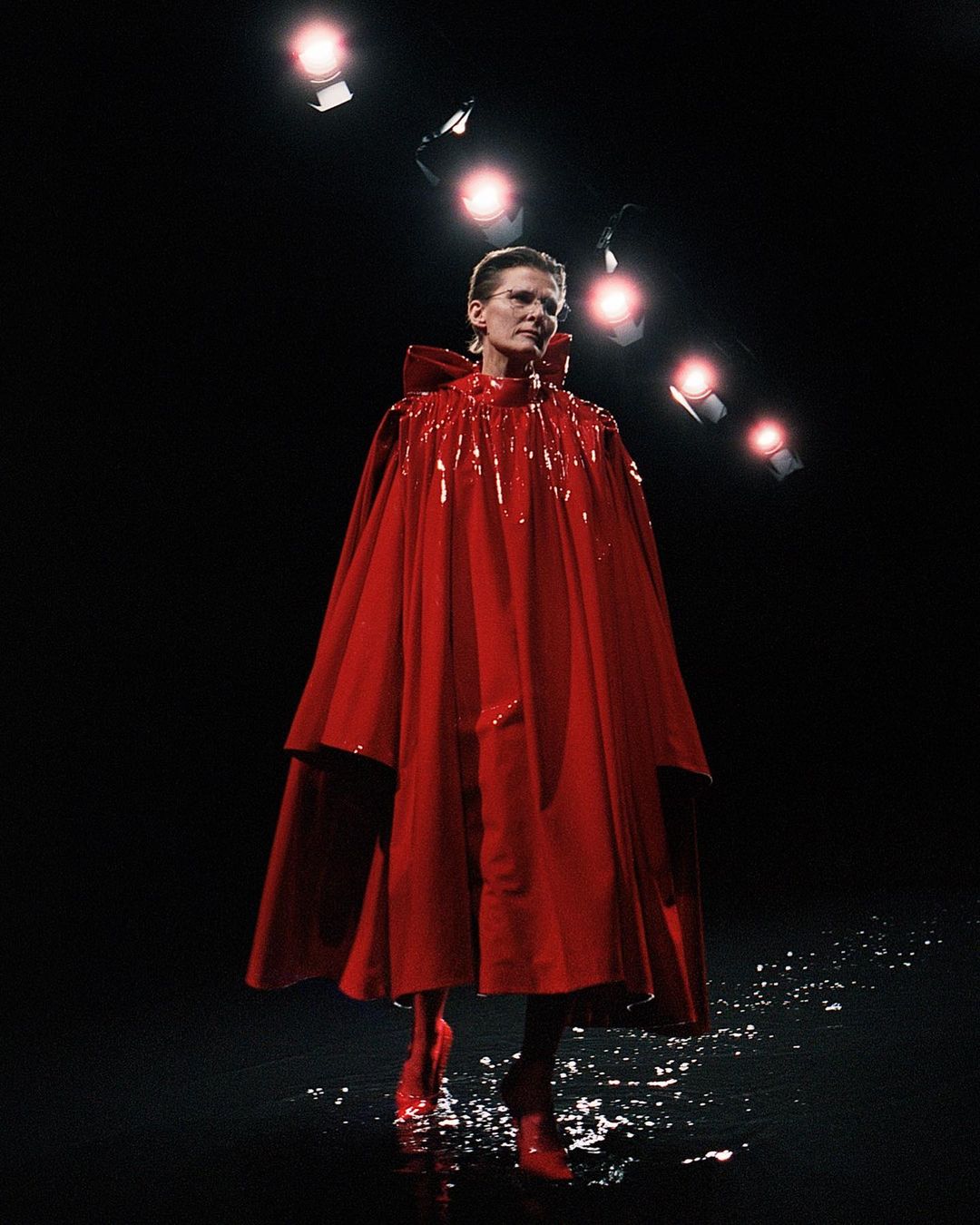 Fashion model wearing a shiny red cape and matching boots on a wet stage under bright stage lights. Dark background in runway show.