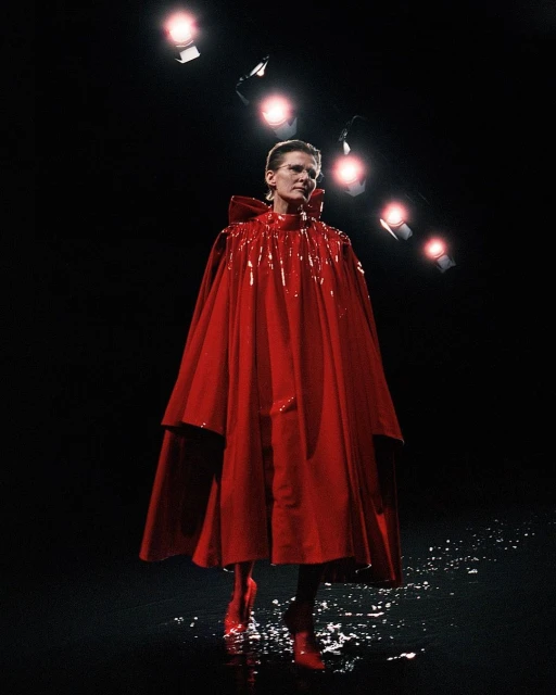 Fashion model wearing a shiny red cape and matching boots on a wet stage under bright stage lights. Dark background in runway show.