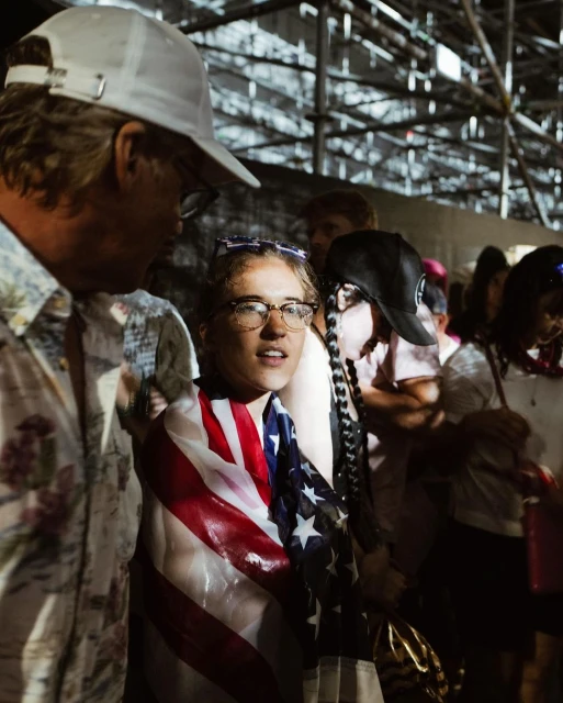 Crowd scene shows a young woman wrapped in an American flag, standing next to a man with a white hat, at an outdoor event.
