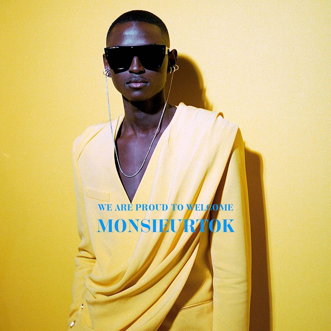 Stylish dark-skinned model wearing a yellow jacket and sunglasses poses against a yellow background, showcasing high fashion and design.